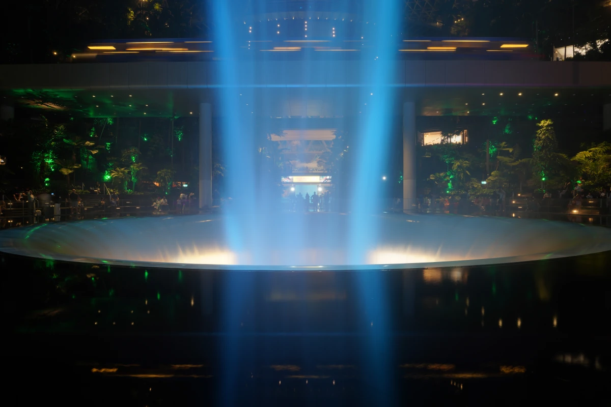 Unteres Becken des Rain Vortex im Jewel Changi Airport in Singapur bei Nacht, im Hintergrund zwei Züge